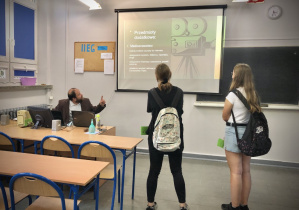 Dwie ósmoklasistki ogladają prezentację o klasie medialnej wyświetlaną na tablicy przez nauczyciela wosu